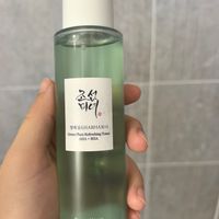 A hand holding a green-tinted bottle of Korean skincare toner with Korean text and "Green Plum Refreshing Toner AHA + BHA" label against a tiled bathroom background