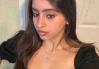 A young woman with long dark hair wearing a black top, taking a selfie indoors