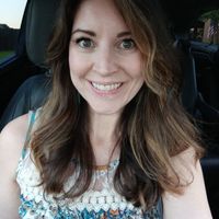A woman with long brown hair smiling while sitting in a car, wearing a floral patterned top with white lace detail
