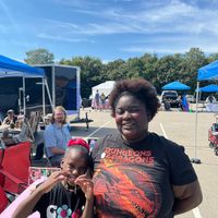 A woman and child pose together at an outdoor event with blue tents and vendors in the background under a partly cloudy sky