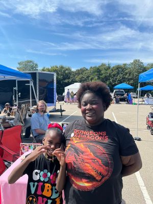 A woman and child pose together at an outdoor event with blue tents and vendors in the background under a partly cloudy sky