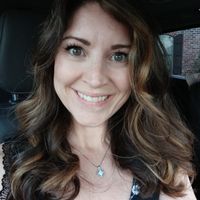 Woman with brown wavy hair and green eyes smiling in a car, wearing a dark floral top and necklace