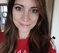 A woman with long brown hair wearing a red t-shirt, smiling at the camera in what appears to be a home setting