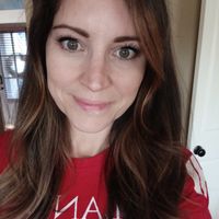 A woman with long brown hair wearing a red t-shirt, smiling at the camera in what appears to be a home setting