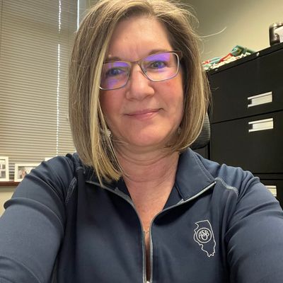 Woman with glasses and blonde hair wearing a navy blue zip-up jacket in an office setting