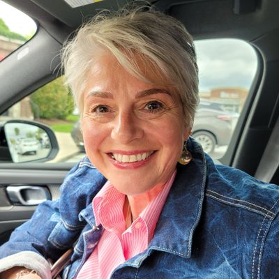 A smiling woman with short blonde hair wearing a denim jacket and pink striped shirt sitting in the driver's seat of a car