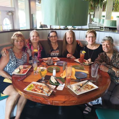 Six women sitting together at a round wooden table in a restaurant, smiling and enjoying a meal with drinks and plates of food