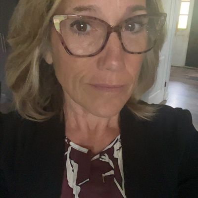 A woman with blonde hair wearing glasses, a dark blazer, and a patterned blouse taking a selfie indoors