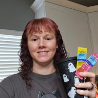 A person with curly reddish hair wearing a gray t-shirt with a cat design, holding colorful hydration drink packets while standing in a kitchen