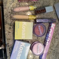Collection of Kosas brand cosmetic products including lip glosses, concealers, blush compacts and other makeup items arranged on a speckled granite countertop