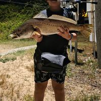 A young person holding a large fish they caught while standing near fishing equipment and a boat mast sign